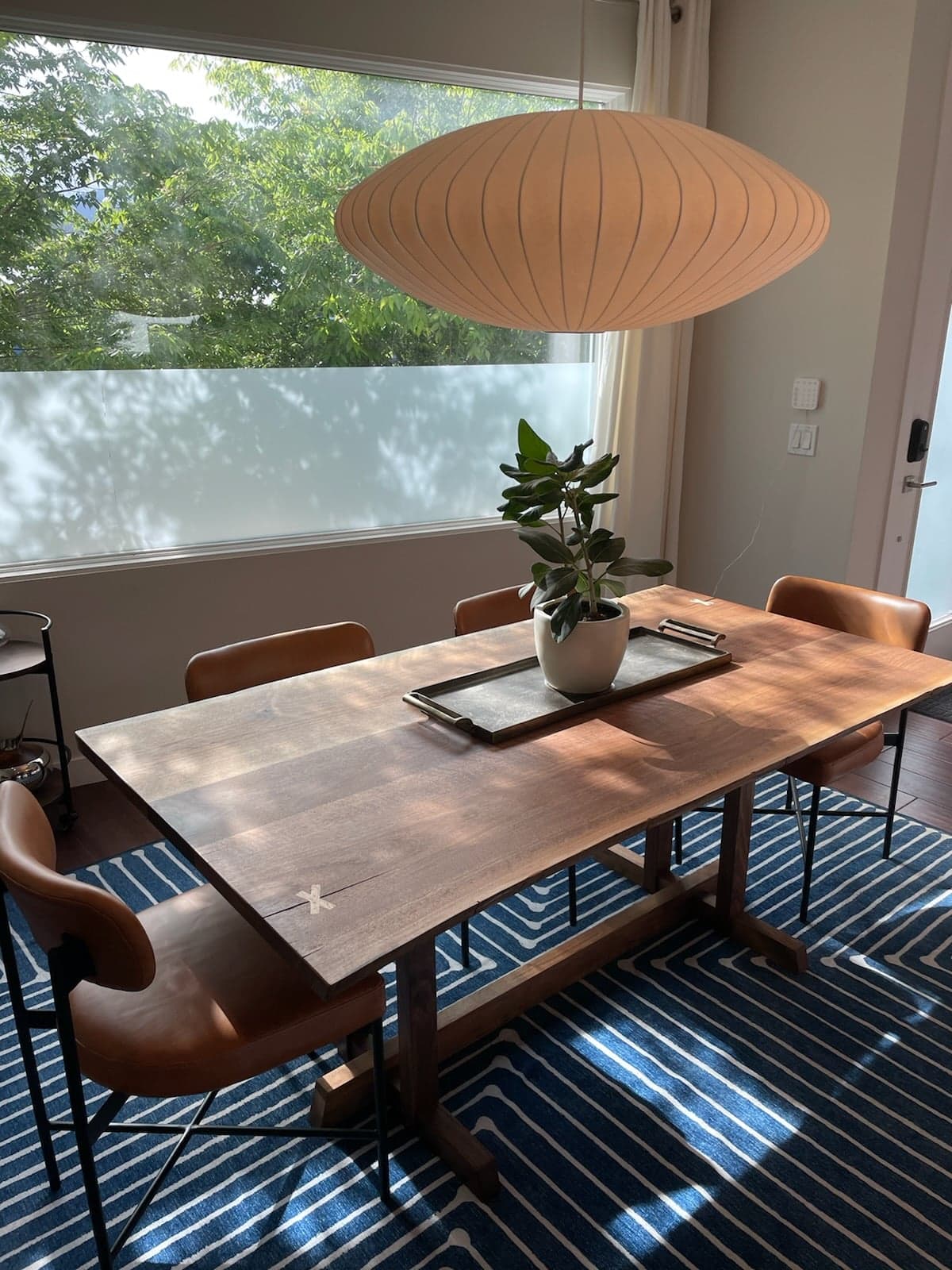 View of table in a modern dining room. The table features several slabs of wood with live edges joined together and x inlays in opposite corners. The trestle base is offset from the edges to ensure a more comfortable sit.