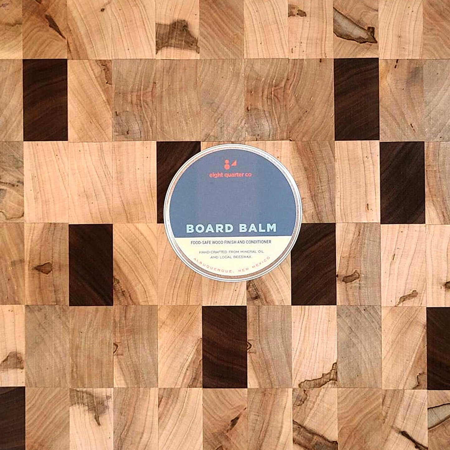 Aerial view of Board Balm tin resting on a wooden surface. The wooden surface consists of rectangles of different colors and types of wood.