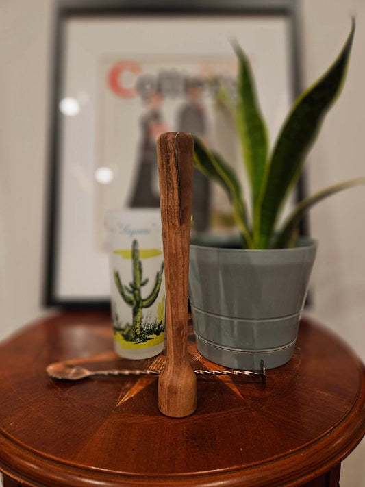 Wooden muddler with a cylindrical bottom and slowly tapering handle in the medium brown color of eucalyptus in focus. It's resting on a small round table with a modern stirring spoon, a potted snake plant, a shot glass with a cactus on it, and framed art behind it.