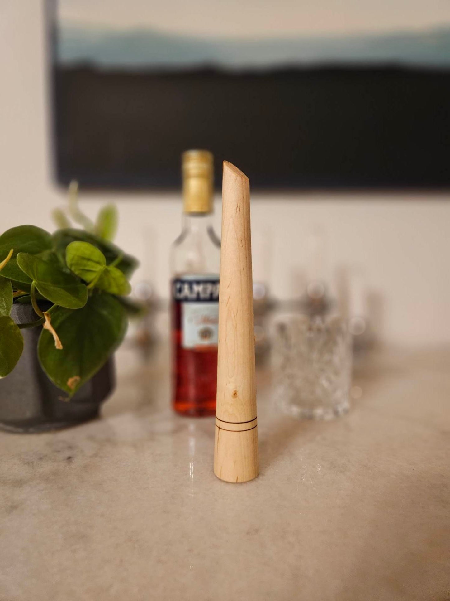 Closeup of a wooden muddler in a light wood color on a counter with cocktail items blurred behind it. The top gently tapers toward the bottom, where 2 thin lines are carved into the circumference. The top is cut at an angle.